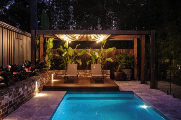 A moody, landscaped outdoor pool area with a compact rectangular swimming pool in the foreground, surrounded by stone or tiled paving. Behind it is a timber deck with two lounge chairs, dense tropical planting, brick retaining walls, and a dark timber pergola with a slatted ceiling and integrated lighting. A timber-clad structure and glass fencing frame the secluded courtyard-like setting.
