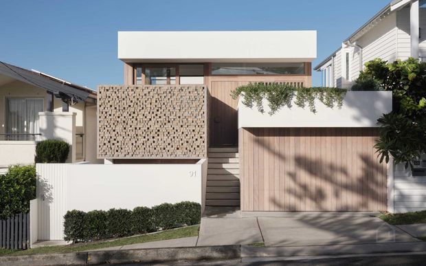 Street-facing exterior of a compact contemporary multi-level residence with a crisp white box-like upper volume, warm timber cladding and garage door, a patterned breeze-block privacy screen, and landscaped entry planting. The home sits in a dense suburban streetscape with a front stair and integrated garage.