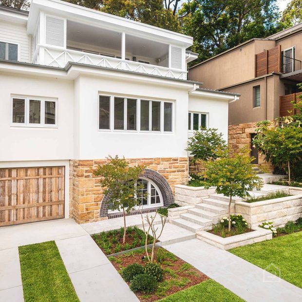 Exterior view of a refined two-storey residence with a white rendered facade, covered upper-level veranda, decorative balustrade, tall white-framed windows, timber garage door, and a sandstone-clad lower level. Landscaped front garden paths and stepped masonry planting beds lead to the entrance, with a neighbouring contemporary residence visible at right.
