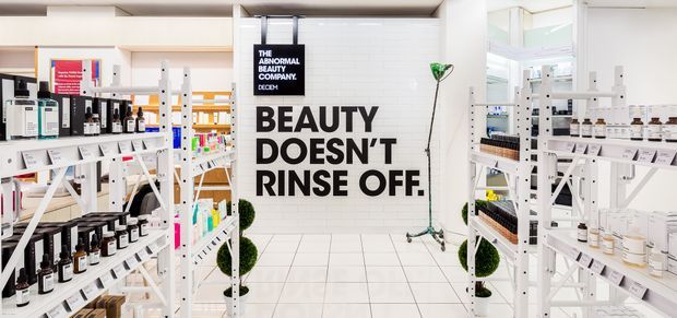 Bright, clean retail beauty showroom with white shelving densely stocked with skincare and cosmetic products. A central tiled wall carries bold black promotional typography, while white tiled flooring, pale wood shelving accents, and strong daylight create a crisp contemporary commercial interior.