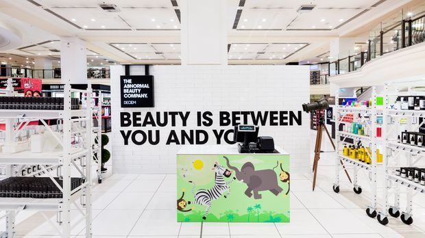 Bright, spacious modern retail beauty store interior with white tiled floors and walls, extensive white display shelving, a central checkout counter with a colourful illustrated animal graphic, black-and-white promotional wall lettering, glass-fronted shelving, and a highly illuminated suspended ceiling.