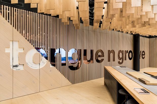 A refined commercial interior reception or showroom with extensive light timber finishes. The space features a sculptural ceiling installation of suspended rectangular timber blocks, vertical timber battens forming a feature wall, integrated timber wall panels, and a large black-and-timber reception or display counter. Bold dimensional lettering is mounted on the batten wall, with glimpses of seating visible behind it.