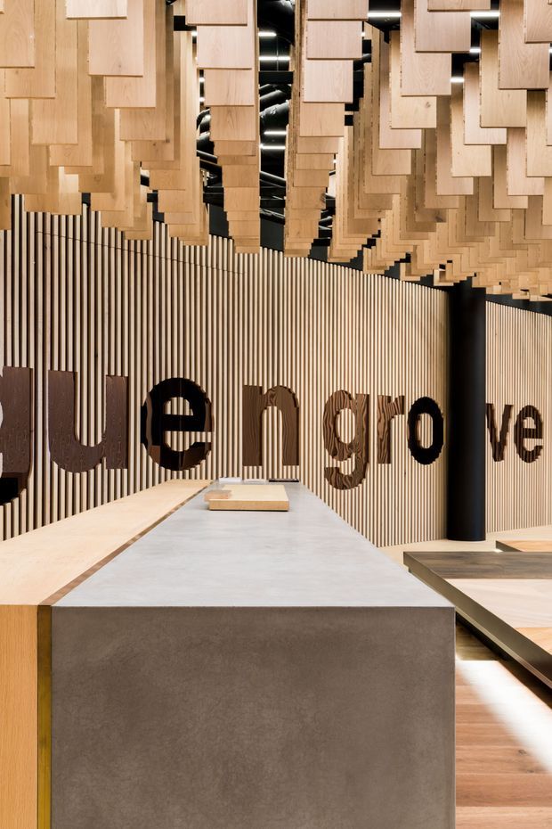 A sophisticated commercial interior with a long central concrete or stone display counter, warm timber flooring, vertical timber slat wall panelling, and large dark lettering forming part of a brand or wayfinding installation. The ceiling features a dramatic suspended composition of staggered timber panels, creating a sculptural canopy over the space. Black cylindrical structural or display columns and dark ceiling elements add contrast. The image appears to show a showroom, reception, or branded retail environment with a refined, contemporary material palette.