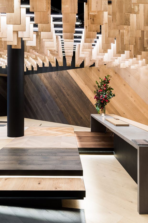 A dramatic contemporary commercial interior or showroom featuring extensive timber flooring displays and a striking suspended ceiling installation made from staggered timber planks. The space combines pale and dark wood surfaces with matte black structural columns, dark joinery, integrated linear lighting, and a long light-coloured counter or display bench. A small floral arrangement adds colour near the counter. The composition is highly geometric, layered, and material-focused.