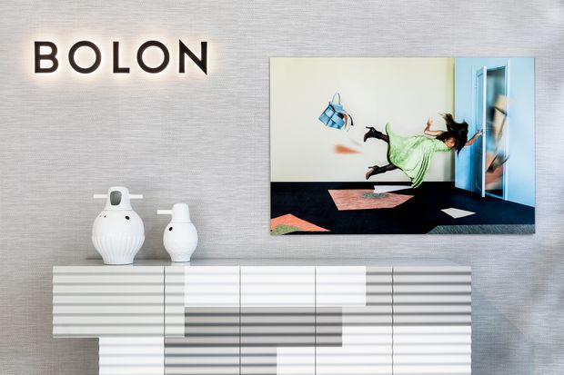 A contemporary showroom-style interior featuring a textured grey wall with illuminated BOLON signage, a large framed artwork or display screen, sculptural white ceramic vessels, and a white linear sideboard with layered horizontal detailing.