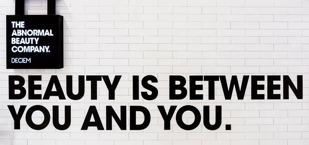A bold typographic wall installation featuring large black lettering reading “BEAUTY IS BETWEEN YOU AND YOU.” on a white painted brick wall, with a small black sign above reading “THE ABNORMAL BEAUTY COMPANY. DECIEM.” The image is graphic, monochromatic, and commercial in character.