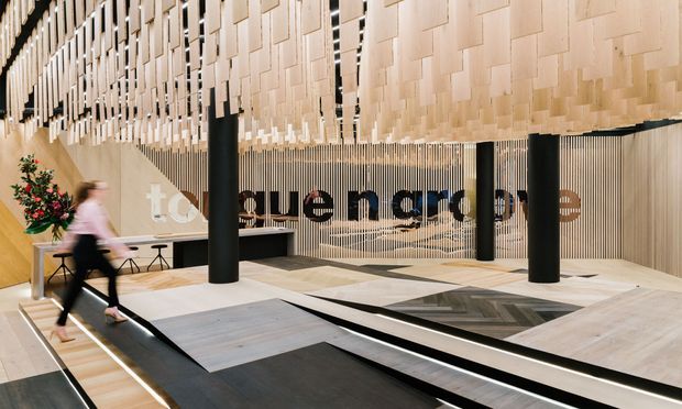 A contemporary commercial interior or public lobby featuring a dramatic suspended ceiling installation of layered light timber or wood-toned panels, black cylindrical columns, pale timber wall cladding, and a geometric floor with contrasting dark bands and inset glazed sections. A large typographic wall graphic and a few furnishings are visible, creating a refined, gallery-like reception or exhibition environment.