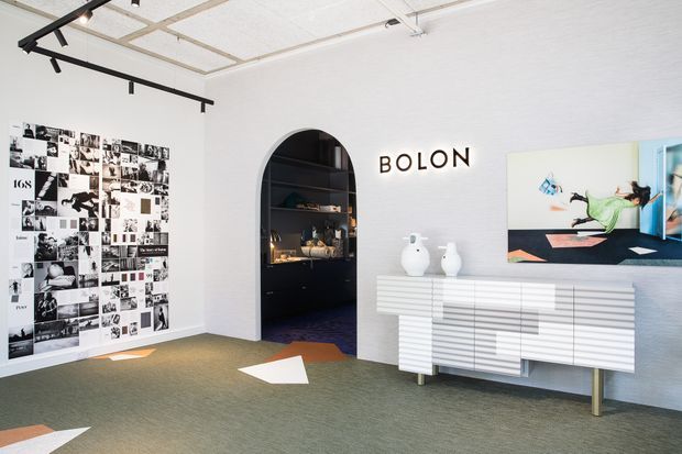 Bright, minimalist commercial showroom or gallery interior with white textured walls, a distinctive arched black display niche, white ribbed console cabinetry, track lighting, grey-green carpet, monochrome wall collage, and contemporary artwork. The space has a clean, refined retail or design-studio presentation.
