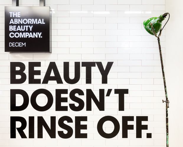 A graphic retail or commercial interior feature wall clad in white rectangular subway tiles. Large black typographic lettering reads “BEAUTY DOESN’T RINSE OFF,” with a black framed DECIEM sign mounted at upper left and a green industrial-style wall lamp at right. The composition is bold, monochromatic, and highly graphic.