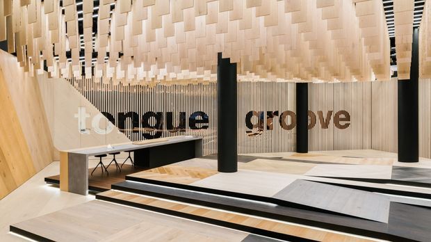 A contemporary commercial interior or exhibition space featuring extensive light timber wall and ceiling treatments, a sculptural suspended ceiling of overlapping rectangular panels, vertical timber battens, black cylindrical columns, and stepped geometric platforms in black, white, and pale wood. A long communal table and seating are visible in the background, with large “tongue groove” lettering integrated into the display wall.