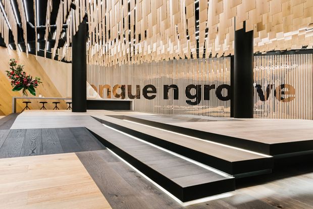 Contemporary commercial interior featuring a dramatic stepped platform or stage, broad timber flooring, black structural columns, and a highly distinctive suspended timber ceiling and slatted feature wall. Large dark lettering is integrated into the rear screen, while linear lighting emphasizes the steps and ceiling. A small seating or hospitality area is visible to the left.