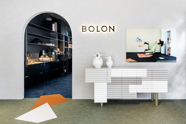 A refined modern living room or showroom vignette featuring a sculptural white ribbed sideboard, wall-mounted television, ceramic vessels, and a dramatic arched opening into dark built-in shelving. The space has textured light walls, muted green-grey carpet, and geometric orange and white floor accents.