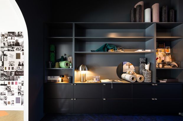 A dark, sophisticated built-in storage and display wall dominates the room, with matte black cabinetry, open grey shelving, integrated lighting, and curated books, ceramics, textiles, and decorative objects. A large monochrome graphic artwork is partially visible at the left edge, and the cabinetry sits above a dark patterned floor.