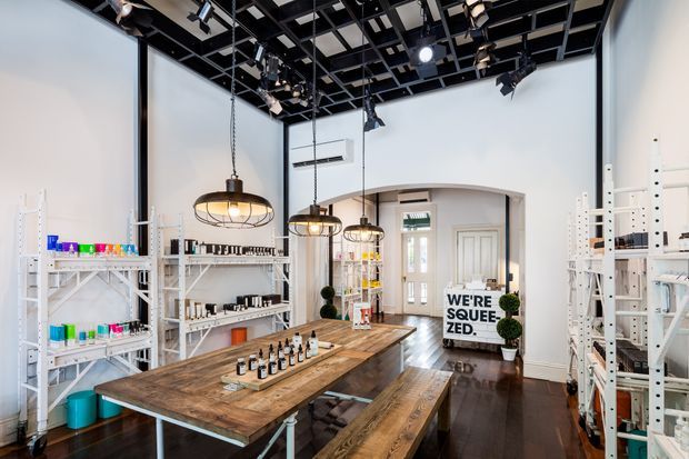 Bright retail showroom or studio with white shelving displaying small products, a large rustic timber worktable and benches, pendant lights, exposed black ceiling grid with track lighting, and a small entry area at the rear. The space has a creative, industrial-inspired fit-out with white walls and dark timber flooring.