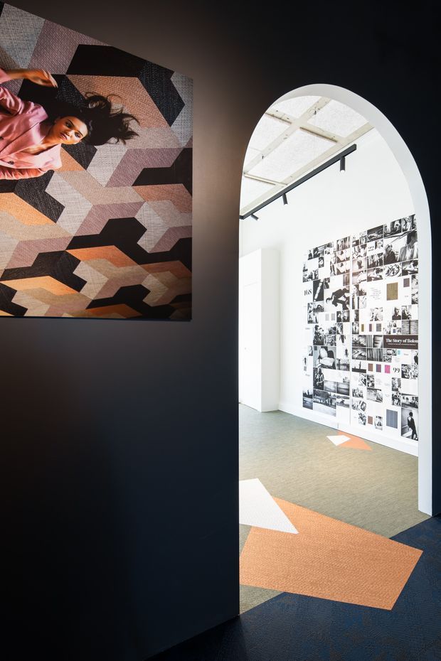 A contemporary commercial interior viewed through a tall arched opening. A dark charcoal wall in the foreground displays a large geometric abstract artwork, while the bright adjoining space features white walls, exposed ceiling structure, linear track lighting, a large black-and-white photographic collage, and a patterned carpet in orange, cream, grey, and navy tones.
