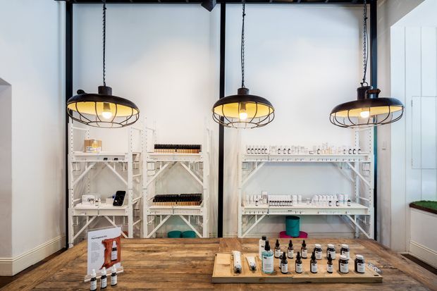 A bright retail display space with industrial-style pendant lighting, white open shelving stocked with small products, and a large rustic timber display table in the foreground. Black-framed full-height glazing or partitions and white walls create a clean, contemporary shop interior.