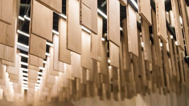 A close-up view down a dramatic interior installation featuring numerous vertically suspended timber panels arranged in repeating rows, with linear strip lighting and small spotlights illuminating the warm wood surfaces. The space has a sculptural, immersive and possibly commercial exhibition-like character.