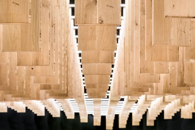 A highly symmetrical architectural interior detail featuring repeated suspended or projecting light timber panels and slatted elements arranged in a long corridor-like composition. The warm natural wood creates a rhythmic, geometric ceiling or canopy installation, with dark negative space visible beyond.