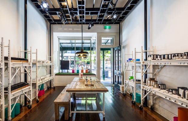 A bright, narrow retail showroom or boutique shop with white walls, dark timber flooring, open pale timber shelving displaying packaged products, and a central timber worktable with stools. Large front windows and a glazed entrance provide views toward the street. The ceiling has exposed black services, track lighting, and suspended fixtures, creating an industrial-style fit-out.