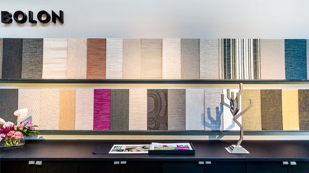 A Bolon showroom or display installation featuring rows of vertically arranged textured and patterned flooring samples in varied neutral, earth, and accent colours. The samples are mounted on a backlit wall above a dark display counter with brochures, a floral arrangement, and sculptural decor.