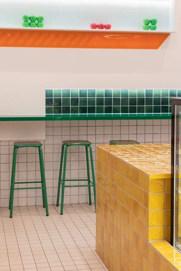A brightly coloured tiled interior, likely a café or commercial bar space. A yellow-orange tiled counter dominates the foreground, with green metal bar stools, pale square wall and floor tiles, a dark green tiled backsplash, and a vivid orange angular overhead shelf or canopy. Green and red decorative objects sit on the upper shelf. The space has a playful, retro-inspired colour scheme and a clean geometric composition.