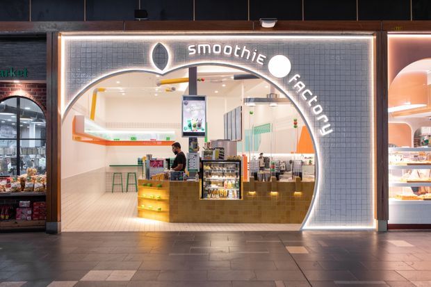 A contemporary smoothie shop storefront in an indoor commercial setting. The facade features a large illuminated arched opening, dark tiled framing, a glowing “smoothie factory” sign, and a bright interior with tiled walls, a service counter, display refrigerators, stools, and food shelving.