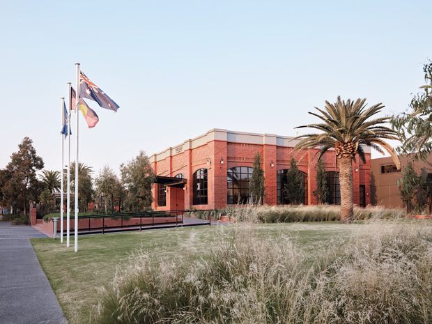 Exterior view of a low-rise red brick institutional or civic building with tall arched steel-framed windows, landscaped grounds, flagpoles, ornamental grasses, and palm trees. A paved accessible ramp leads toward the entrance. The architecture has a warm, formal Mediterranean-influenced character with strong masonry detailing.