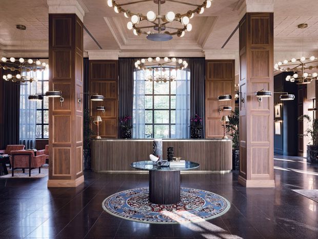 A grand, symmetrical hotel or commercial reception lobby with dark polished flooring, tall timber-panelled columns, a ribbed curved reception desk, black-framed floor-to-ceiling windows, and ornate multi-globe chandeliers. A round decorative rug and central circular table create a formal focal point, while lounge seating and plants sit around the perimeter. The space has a richly detailed, luxurious contemporary-classic character.