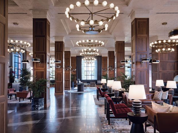 Grand, richly detailed hotel or commercial lobby/lounge with a symmetrical arrangement of dark timber-clad columns, polished dark stone flooring, mirrored walls, ornate coffered ceilings, and repeated circular globe chandeliers. The space includes lounge seating, side tables, lamps, patterned rugs, and large windows at the far end, creating a refined and dramatic atmosphere.