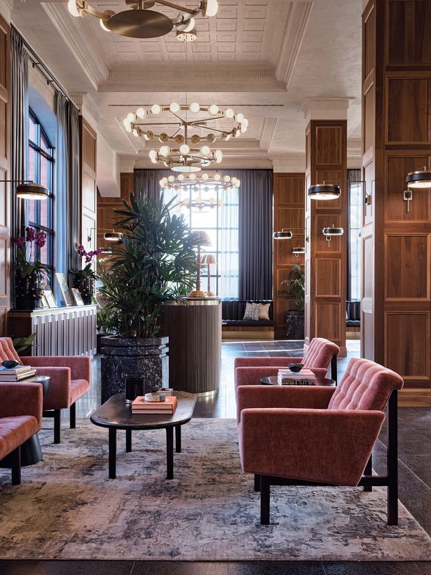 Elegant, richly detailed living room or lounge with tall windows, dark timber wall panelling and built-in cabinetry, ornate coffered ceiling, layered globe chandeliers, and multiple dusty-rose upholstered lounge chairs arranged around a dark oval coffee table. A large patterned rug, indoor plants, decorative objects, and a rear seating area create a sophisticated hospitality-style interior with strong symmetry and abundant natural light.