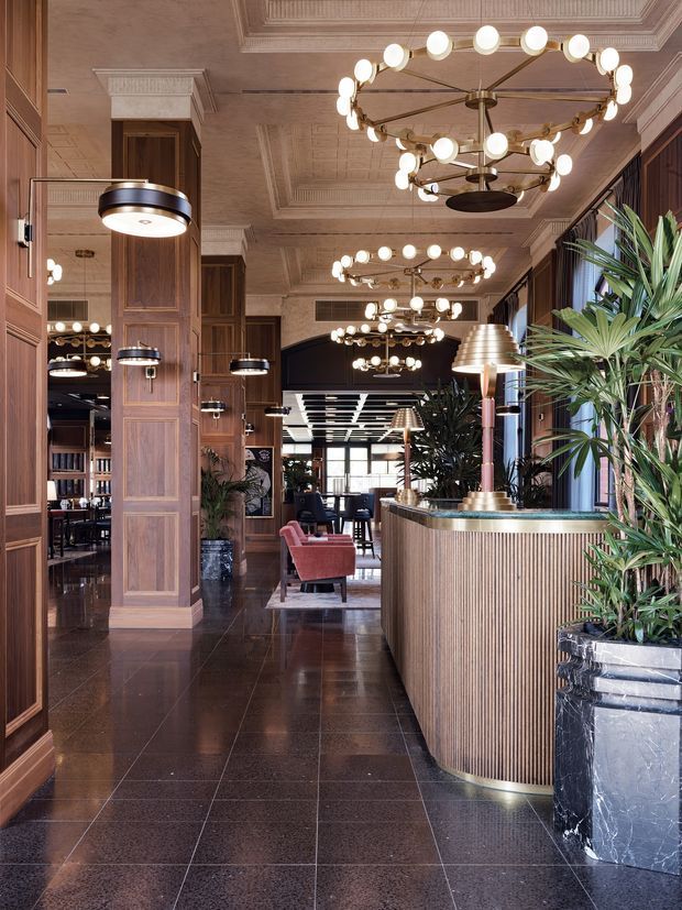 A refined hospitality-style interior, likely a hotel lobby or restaurant reception, with dark polished stone tile flooring, rich timber wall panelling and columns, ornate decorative ceiling detailing, and repeated spherical brass-and-opal pendant chandeliers. A curved fluted timber reception counter, indoor tropical planting, upholstered lounge chairs, mirrors, and warm layered lighting create a luxurious, classic atmosphere.