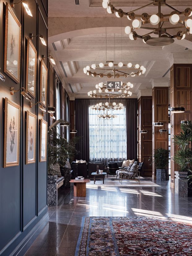 A grand, hotel-like interior lobby or reception hall with a long axial layout, polished dark stone flooring, rich navy-blue paneled walls, dark timber architectural joinery, framed artwork, and tall sheer-curtained glazing at the far end. Multiple sculptural ring chandeliers with globe lights run along the ceiling, while lounge seating, indoor plants, and patterned rugs create a refined hospitality atmosphere. Ornate decorative ceiling detailing and strong symmetry contribute to the luxurious, dramatic character.