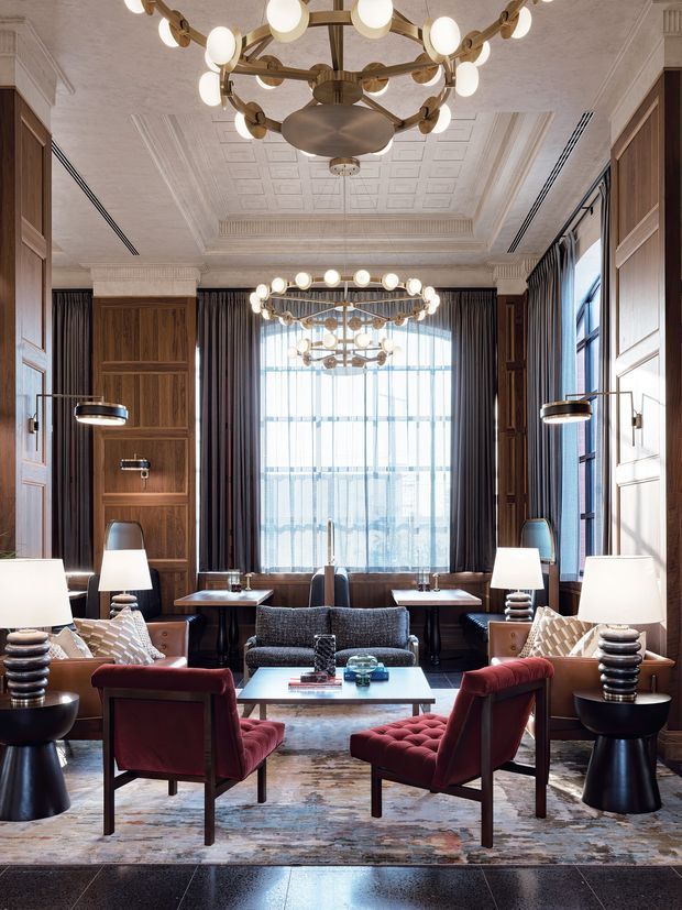An elegant, formal lounge or hotel-style sitting room with richly detailed dark timber wall panelling, ornate coffered ceiling, classical cornice and pilaster detailing, tall windows with dark curtains, and multiple sculptural brass-and-glass chandeliers. The room is furnished with burgundy velvet armchairs, a dark sofa, side tables, table lamps, and a central glass-topped coffee table over a large patterned rug. The scheme is luxurious, refined, and traditional with contemporary furnishings.