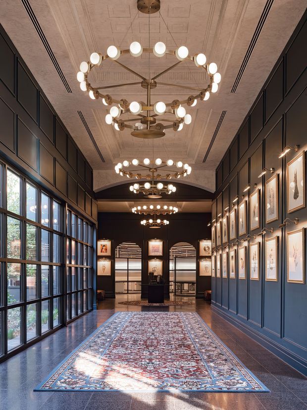 A grand, elongated interior hallway or entry gallery with dark blue-black paneled walls, tall multi-pane windows on one side, polished dark stone flooring, and a richly patterned runner rug. The space features a coffered and ornamented ceiling with multiple large circular brass-and-globe chandeliers, framed artwork arranged along the walls, and a view toward a formal reception or lounge area at the far end. The overall character is elegant, dramatic, and Art Deco-inspired.
