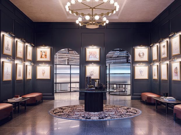 Elegant, symmetrical gallery-like interior lobby with deep navy panelled walls displaying framed artwork, arched glass openings, polished stone flooring, a central dark pedestal table, burgundy upholstered benches, and an ornate circular chandelier. The space has a refined, luxurious feel with warm brass and cream accents.