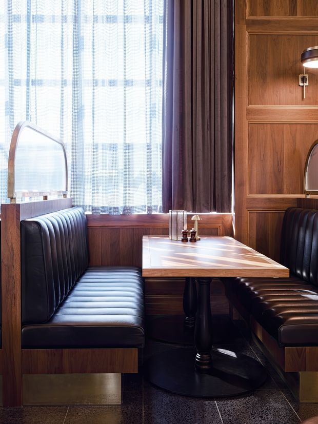 A richly detailed dining booth with dark leather banquette seating, a polished timber pedestal table, timber wall panelling, and a large window dressed with sheer and dark curtains. Brass-toned plinths and hardware, a vintage-style wall light, and a dark tiled floor create a warm, refined restaurant or hospitality interior with strong Art Deco influence.