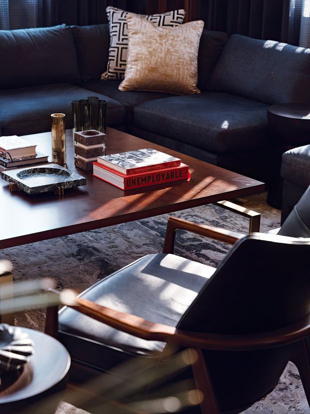 A moody, refined living room with a large dark charcoal sectional sofa, patterned and textured cushions, a low rectangular timber coffee table, decorative vessels, books and a prominent red book, plus a dark leather-and-timber lounge chair in the foreground. A patterned grey rug anchors the seating area, while strong sunlight creates dramatic highlights and shadows through the windows.
