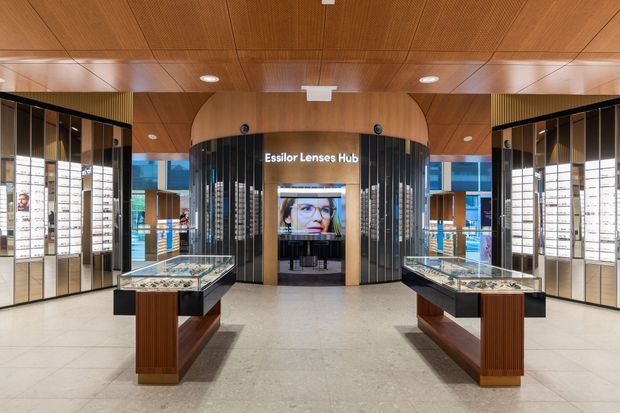 A symmetrical contemporary optical retail showroom interior featuring illuminated wall displays, glass-topped product islands, a central curved Essilor Lenses Hub, dark vertical slatted detailing, and extensive warm timber ceiling panelling. The polished light floor and bright integrated lighting create a refined commercial environment.