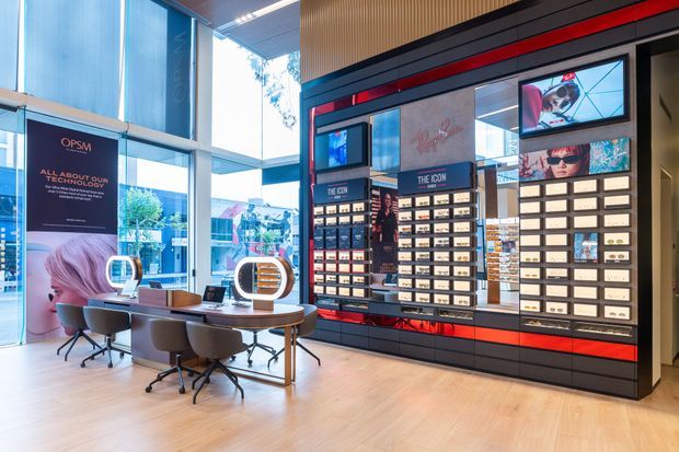A bright contemporary optical retail showroom with a consultation table and rolling chairs in the foreground, large glazed windows, and a dramatic black, red, and timber-toned product display wall featuring illuminated shelving and digital screens.