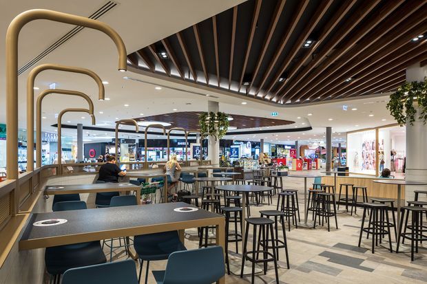 A contemporary indoor shopping-centre dining and food-court area with numerous communal tables, bar-height stools, booth seating, and integrated charging points. The space features a dramatic timber slatted ceiling with recessed lighting, curved brass-toned overhead frames, pale flooring, planted columns, and retail storefronts visible in the background.