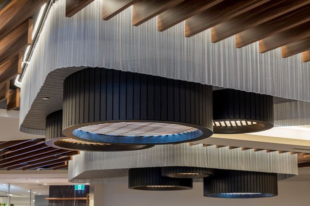 Interior architectural view of a contemporary commercial space featuring a dramatic ceiling installation. Large dark ribbed cylindrical pendant forms hang beneath a patterned ceiling of vertical translucent fins, with exposed warm timber beams and integrated linear lighting. The composition emphasizes layered ceiling geometry and sculptural lighting rather than furnishings.