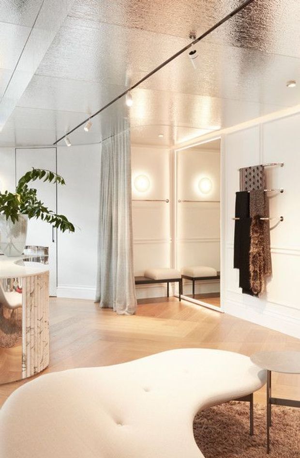 Bright, refined interior featuring a sculptural cream upholstered seating piece in the foreground, a mirrored cylindrical side table, pale herringbone timber flooring, full-height mirrors, sheer curtains, built-in wall mouldings, and a wall-mounted towel or textile rack. Track and recessed lighting create a warm, polished atmosphere, with a large indoor plant adding an organic touch.