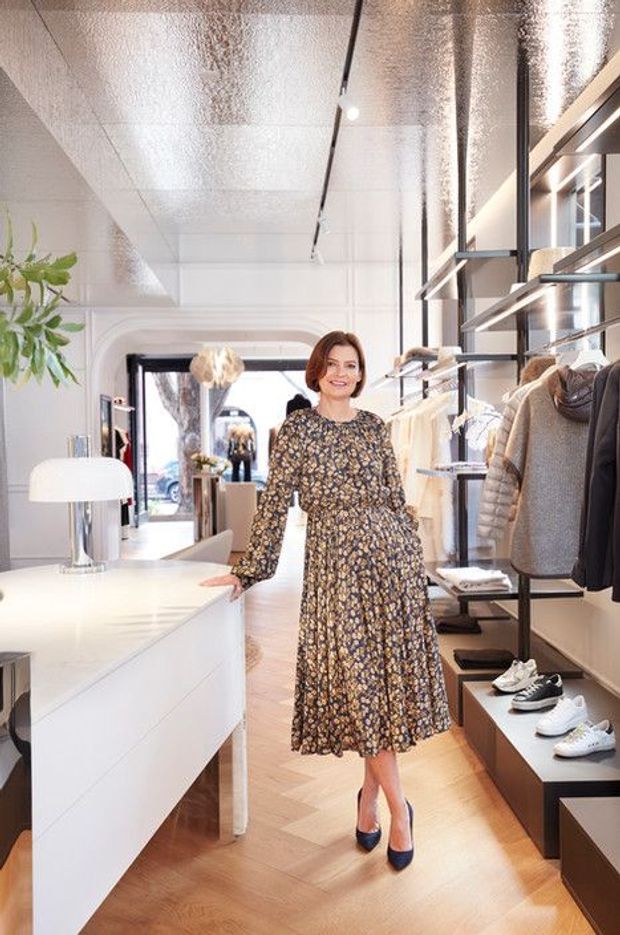 Bright, modern retail clothing boutique interior with a woman standing beside a white display counter. The space features clothing racks and shelving, shoes displayed on dark plinths, warm timber parquet flooring, a reflective metallic ceiling, large glazing at the entrance, and contemporary linear lighting. White cabinetry and walls create a clean, refined commercial fit-out.