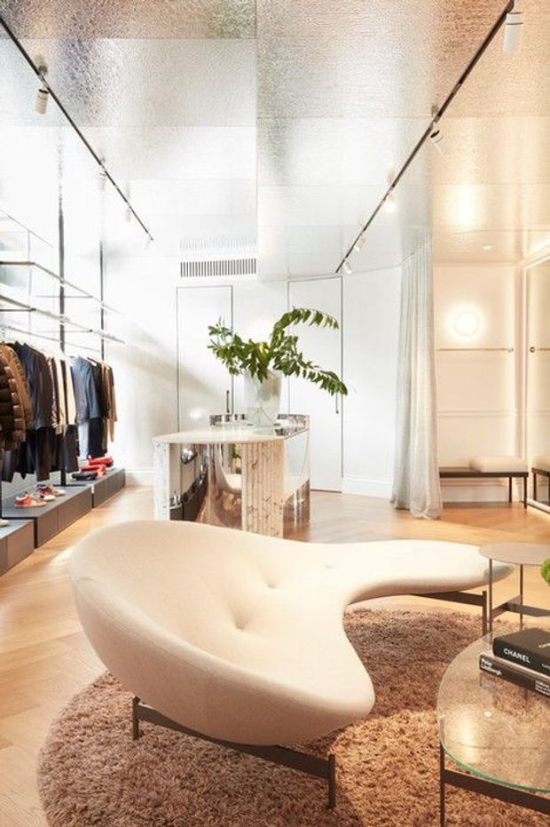 A bright, refined retail-style dressing and lounge space with clothing displayed along built-in shelving, a sculptural cream curved sofa, shag rug, glass coffee table, mirrored central island, and indoor greenery. The room has white walls and ceiling, pale timber flooring, track lighting, and soft ambient lighting, creating a luxurious contemporary boutique atmosphere.