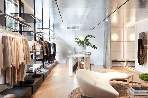 Bright, refined contemporary fashion boutique interior with clothing displayed on open shelving and hanging rails along the left wall. The space features pale timber flooring, white paneled walls, full-height sheer curtains, track lighting, a sculptural cream curved sofa, low tables, a mirrored display plinth, and minimalist styling.