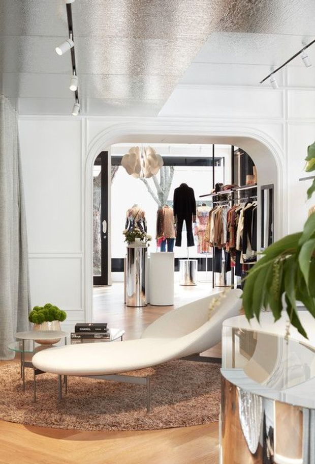 Bright, refined retail interior viewed from a lounge-like foreground area, featuring white decorative wall panelling, an arched opening, track lighting, pale timber flooring, a curved cream upholstered chaise, reflective metallic side tables, a textured rug, plants, and clothing displays in the adjoining space.