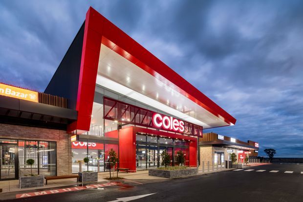 Exterior dusk view of a contemporary Coles supermarket within a retail shopping centre. The building features a bold red projecting entrance canopy and fascia, extensive glass storefronts, illuminated signage, brick and rendered wall sections, timber-toned vertical screening, and a broad asphalt parking area in the foreground beneath a cloudy evening sky.