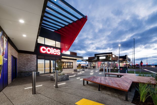Exterior dusk view of a contemporary Coles supermarket and retail centre forecourt. The image shows a large red angular canopy with exposed blue-toned roof glazing or slats, illuminated signage, brick and dark cladding, extensive paved pedestrian areas, timber-topped bench seating, bollards, landscaped planters, and additional low-rise retail buildings beyond. The setting appears commercial and urban, with a road visible in the background.
