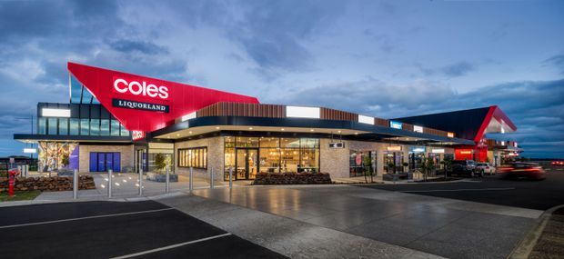 Exterior dusk view of a contemporary Coles supermarket and Liquorland retail complex. The building has a broad low-slung form with glazed retail frontage, dark horizontal fascia, warm timber-look cladding, stone-like wall panels, prominent red angular signage and canopy elements, and a large paved car park in front. The image is primarily about the commercial building exterior.