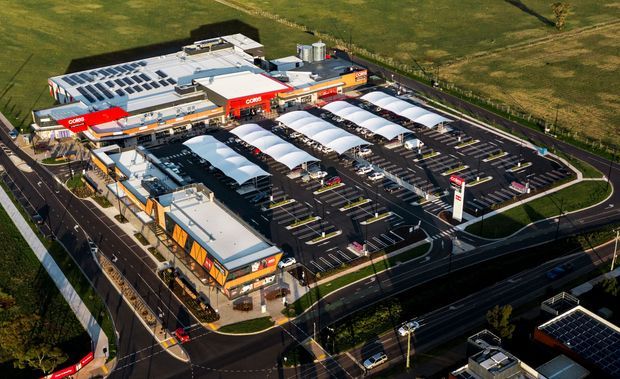 Aerial view of a large commercial shopping centre or retail complex, with multiple big-box buildings, prominent red supermarket signage, extensive parking, covered pedestrian parking canopies, internal access roads, and surrounding open grassland. The image is an exterior site overview rather than an interior space.